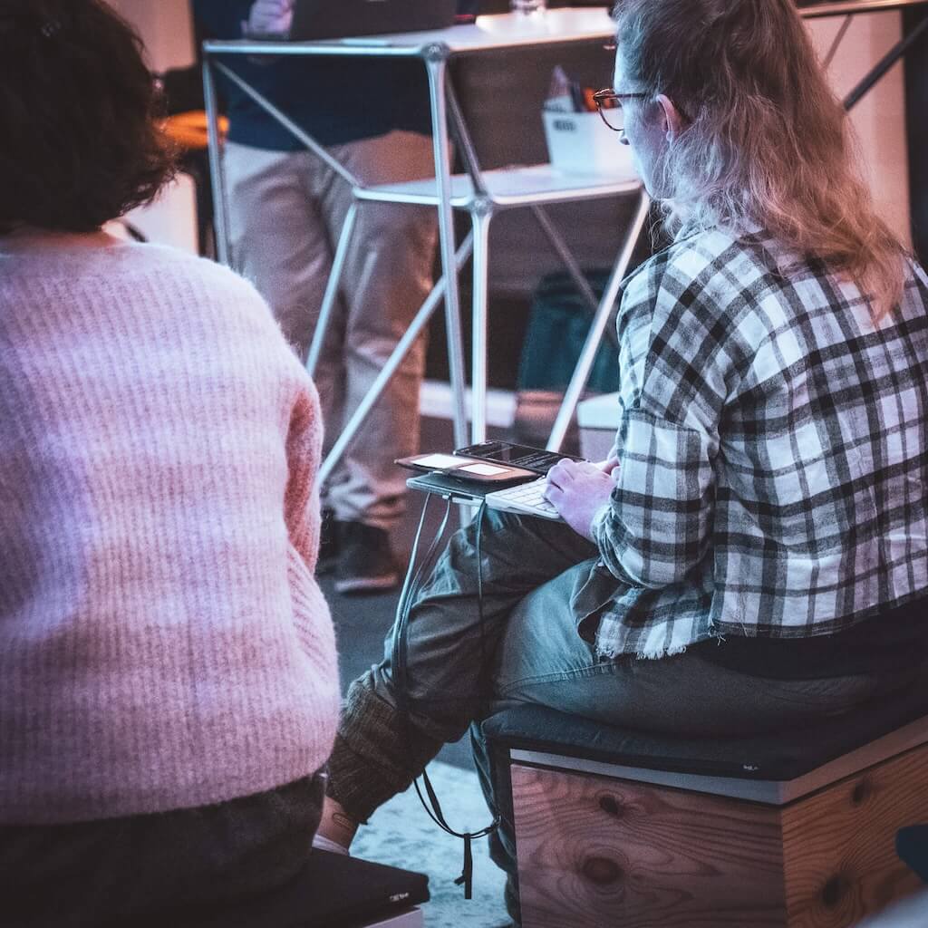 Two people sit on low seats indoors; one person wears a plaid shirt and is taking notes on a tablet, while another sits nearby in a pink sweater. The presenter stands in the background.