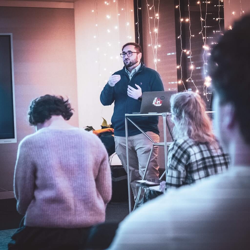 A man stands at a podium giving a presentation to a seated audience in a room decorated with string lights.